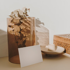 Blank paper sheet card with mockup copy space and dry eucalyptus branch in tan glass vase in sunlight shadows on beige background. Minimal business brand template