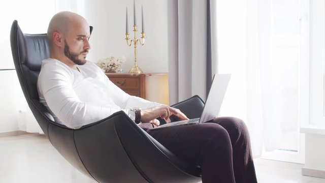 Bearded businessman working at home sitting in armchair and using computer technologies. Business, freelance and stock market concepts.