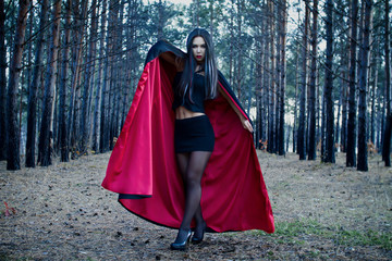 Naklejka premium Vampire Woman in a dark gloomy forest. Beauty Sexy Vampire Girl in a cloak. Sexy Witch. Wicked look, dark hair and red lips. Attractive model girl in Halloween costume and makeup.