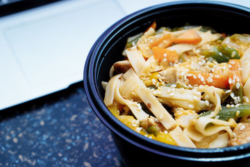 Stir fry noodles with vegetables and chicken in black bowl. Stone background. Top view. Copy space.