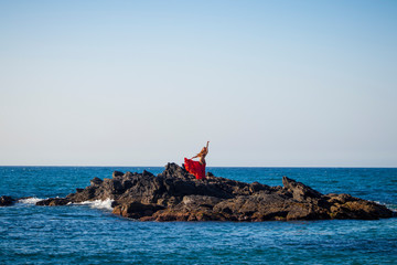 Woman in a red belly dance dress full of ornaments posing on the beach with the rocks and the sea in the background