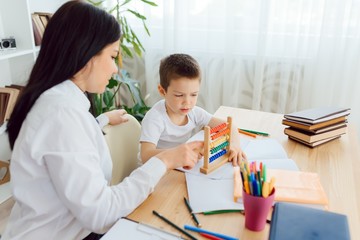 Female private tutor helping young student with homework at desk in bright child's room