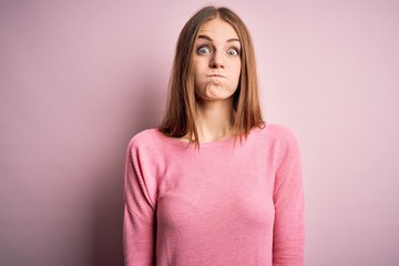 Obraz premium Young beautiful redhead woman wearing casual sweater over isolated pink background puffing cheeks with funny face. Mouth inflated with air, crazy expression.