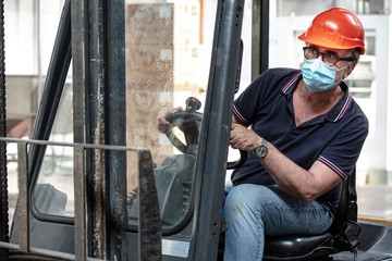 Operaio  con caschetto protettivo e mascherina di protezione guida un carrello elevatore nel magazzino in cui lavora
