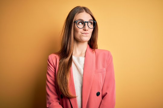 Young beautiful redhead woman wearing jacket and glasses over isolated yellow background smiling looking to the side and staring away thinking.