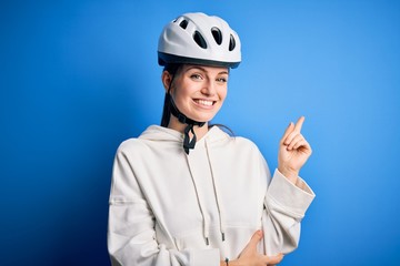 Young beautiful redhead cyclist woman wearing bike helmet over isolated blue background with a big smile on face, pointing with hand and finger to the side looking at the camera.