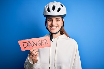 Young beautiful redhead cyclist woman wearing bike helmet holding danger paper message with a happy face standing and smiling with a confident smile showing teeth