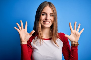 Young beautiful redhead woman wearing casual t-shirt over isolated blue background showing and pointing up with fingers number ten while smiling confident and happy.