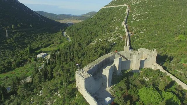 Aerial shot of fort on green mountain against sky, drone is flying over landscape - Ston, Croatia