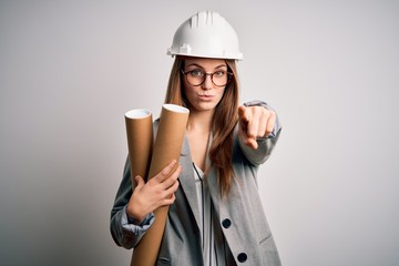 Young beautiful redhead architect woman wearing glasses and safety helmet holding blueprints pointing with finger to the camera and to you, hand sign, positive and confident gesture from the front