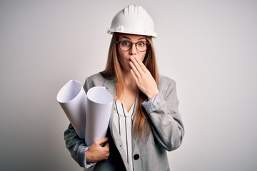 Young beautiful redhead architect woman wearing glasses and safety helmet holding blueprints cover mouth with hand shocked with shame for mistake, expression of fear, scared in silence, secret concept
