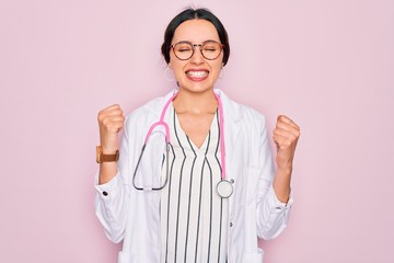 Beautiful doctor woman with blue eyes wearing coat and stethoscope over pink background very happy and excited doing winner gesture with arms raised, smiling and screaming for success. Celebration