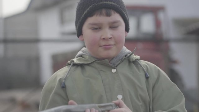 Portrait of cute plump red-cheeked village boy playing with toy gun. Close-up of little charming child with brown eyes resting outdoors on cloudy day. Childhood, leisure, lifestyle.