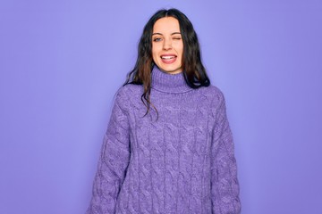 Young beautiful woman wearing casual turtleneck sweater standing over purple background winking...