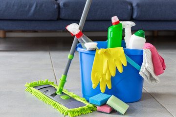 Bucket with bottles of detergent, cleaning tools and mopping stick in front of sofa in living room....
