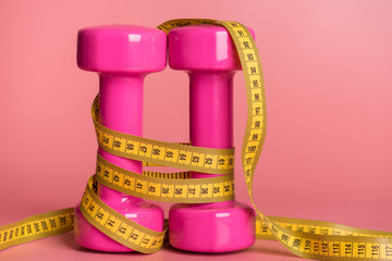 Slimming concept. Close-up photo of two pink dumbbells covered with measuring tape isolated on pastel pink background