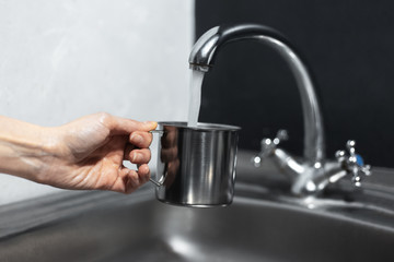 Close-up of female hand holding steel mug under the water tap in kitchen. Background of black and white walls.
