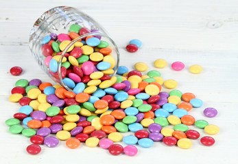 Glass full of colorful and sweet smarties.  Chocolate buttons in colors of rainbow.  Delicious candies on white wooden table.