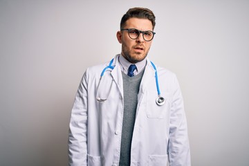 Young doctor man with blue eyes wearing medical coat and stethoscope over isolated background In shock face, looking skeptical and sarcastic, surprised with open mouth
