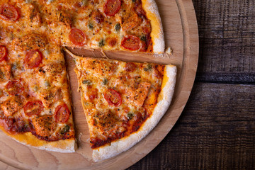 Pizza with salmon, tomatoes and capers on a wooden board. Whole pizza, one piece cut off. Close-up, top view, place for text.