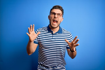 Young man with blue eyes wearing glasses and casual striped t-shirt over blue background crazy and mad shouting and yelling with aggressive expression and arms raised. Frustration concept.