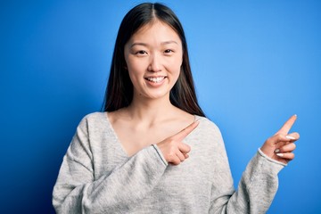 Young beautiful asian woman wearing casual sweater standing over blue isolated background smiling and looking at the camera pointing with two hands and fingers to the side.