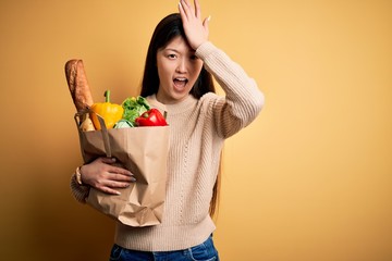 Young asian woman holding paper bag of fresh healthy groceries over yellow isolated background surprised with hand on head for mistake, remember error. Forgot, bad memory concept.