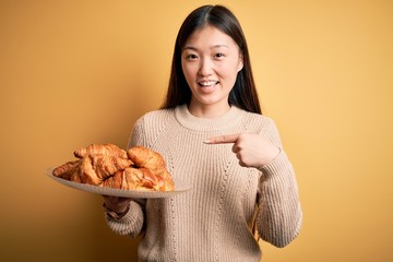 Young asian woman holding french pastry croissant over yellow isolated background very happy pointing with hand and finger