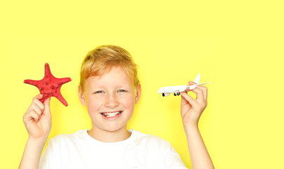Summer vacation concept, travel.Happy smiling 9 year old Caucasian boy i holds a starfish and toy plane in his hands on a yellow background. Kid having fun on summer vacation. Copy space
