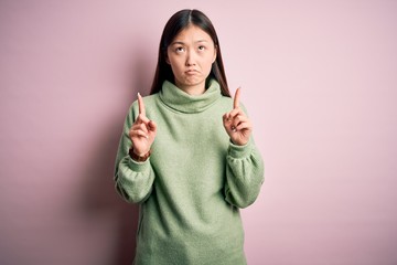 Young beautiful asian woman wearing green winter sweater over pink solated background Pointing up looking sad and upset, indicating direction with fingers, unhappy and depressed.