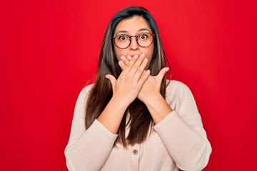 Young hispanic smart woman wearing glasses standing over red isolated background shocked covering mouth with hands for mistake. Secret concept.