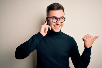 Young handsome caucasian man having a conversation talking on smartphone pointing and showing with thumb up to the side with happy face smiling