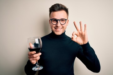 Young handsome caucasian man drinking an alcoholic glass of red wine over isolated background doing ok sign with fingers, excellent symbol