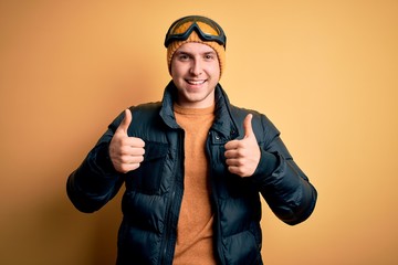 Young handsome caucasian man wearing hat, coat and ski glasses for winter and snow weather success sign doing positive gesture with hand, thumbs up smiling and happy. Cheerful expression and winner