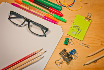 many different stationery and office supplies on a wooden table. black glasses on a pile of white sheets, pens and pencils, paper clips and markers, everything for learning or school