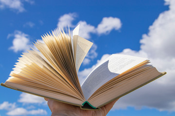 An open book in a hand raised to the sky. Blue sky with clouds create a background for the book.
