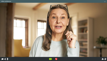Portrait of casually dressed energetic elderly woman with loose gray hair posing in stylish apartment interior looking at camera with mouth opened, speaking via virtual chat, making webcam video call - obrazy, fototapety, plakaty