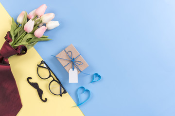 Happy Fathers Day background concept.Decorated red necktie, tulip, blue heart, mustache, eyeglasses, gift box with greeting card on bright pastel background with copy space. Top view, flat lay.