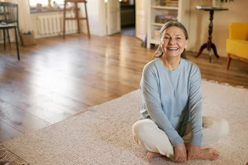 Fotobehang Mature people, active healthy lifestlyle and wellness. Energetic beautiful female pensioner sitting barefooted on carpet keeping legs crossed in half lotus pose, doing morning yoga, smiling © shurkin_son
