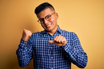 Young handsome latin man wearing casual shirt and glasses over yellow background Pointing to the back behind with hand and thumbs up, smiling confident