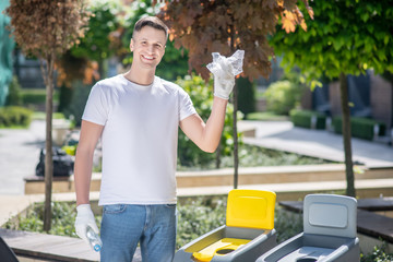 Dark-haired male in protective gloves holding plastic bag in front of waste container