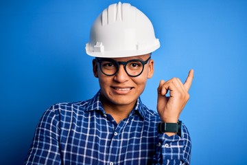 Young handsome engineer latin man wearing safety helmet over isolated blue background with a big smile on face, pointing with hand and finger to the side looking at the camera.