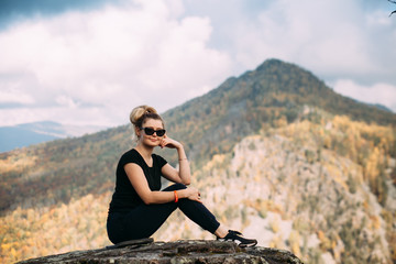 Naklejka na ściany i meble Beautiful russian blondy girl with long hair in sunglasses in black t-shirt sitting on the top of mountain and showing another one 
