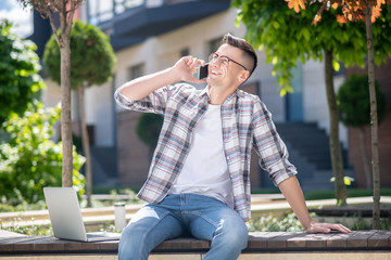 Dark-haired male wearing glasses, sitting on the bench outside with his laptop, talking on his mobile