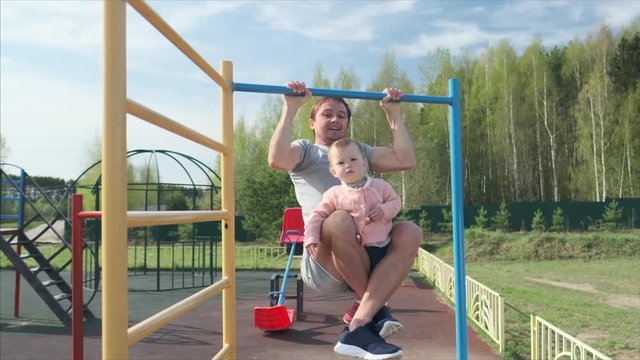 Family work out. Young man does pull ups with his baby. Adult male performs work out with his little child as additional weight.