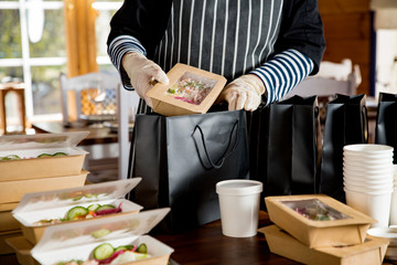 Restaurant worker wearing protective mask and gloves packing food boxed take away. Food delivery services and Online contactless food shopping.