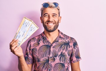 Young handsome blond tourist man with beard on vacation holding airline boarding pass looking positive and happy standing and smiling with a confident smile showing teeth