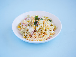 pasta salad plate with corn, cheese and jam, parsley on top, horizontal view on a blue surface with copy space