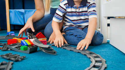 Closeup image of little toddler boy wth young mother assembling toy railroad on floor