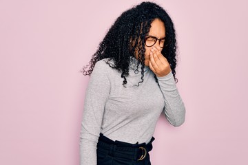 Young african american woman wearing turtleneck sweater and glasses over pink background smelling...